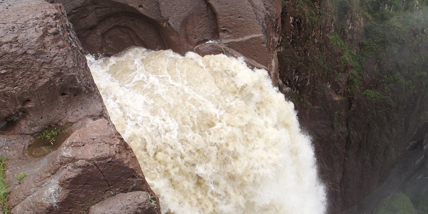 Over a high canyon wall, the mouth of a sharp waterfall cascades into wooded areas.