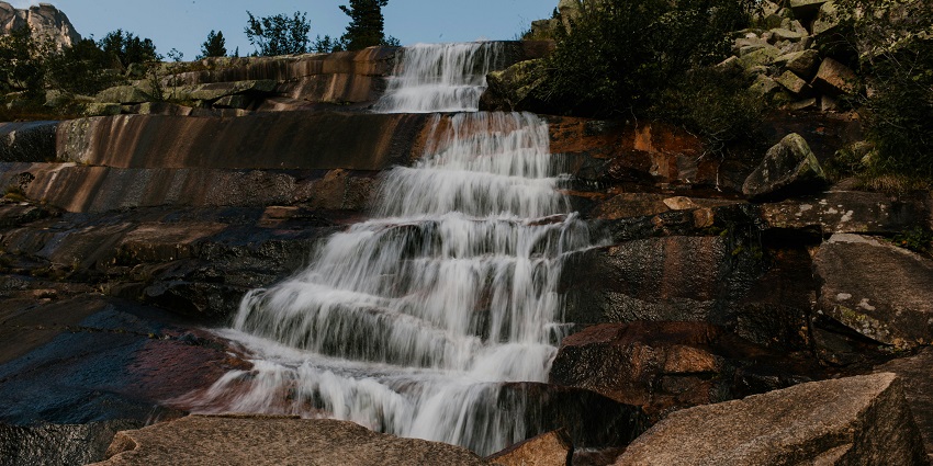 A layered waterfall flows over smooth and dark tiers of rock surrounded by boulders.