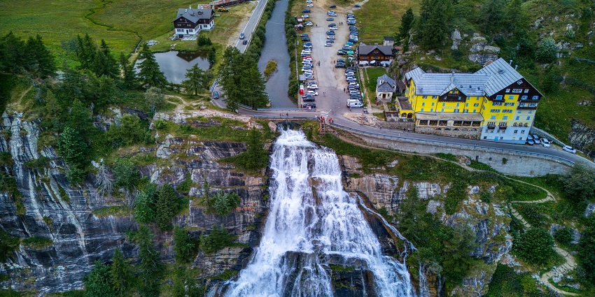 Cascata del Toce flowing from a cliff with a road passing over it, and buildings and cars parked