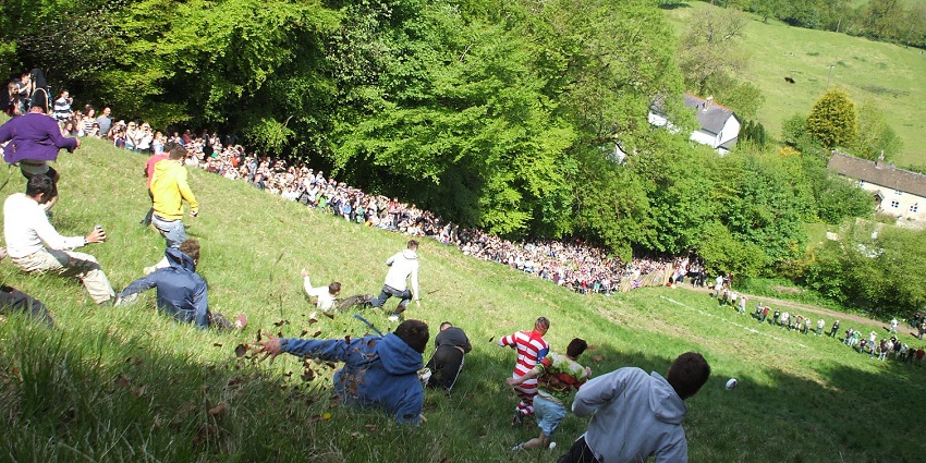 Participants slide along a hill chasing a block of cheese while the audience is gathered at one side