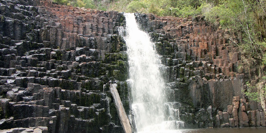 A narrow stream of water flowing over horizontal rows of rock formations into a pool.