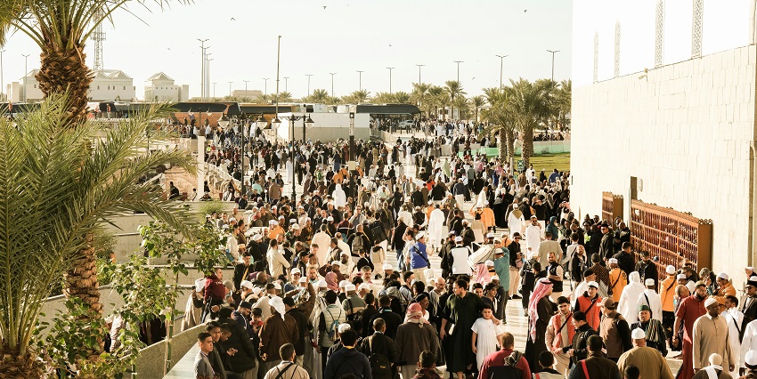 Large crowd in traditional attire walks through a palm-lined area on a sunny day.