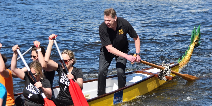 Single dragon boat with a man standing and rowing, others seated and paddling together