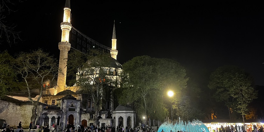 Worshippers praying in a grand, beautifully lit mosque with detailed, ornate interiors.