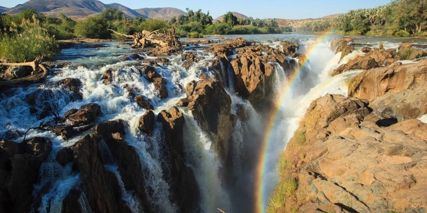 Scenic view of Epupa Falls with water cascading over rocks and a rainbow forming in the mist.