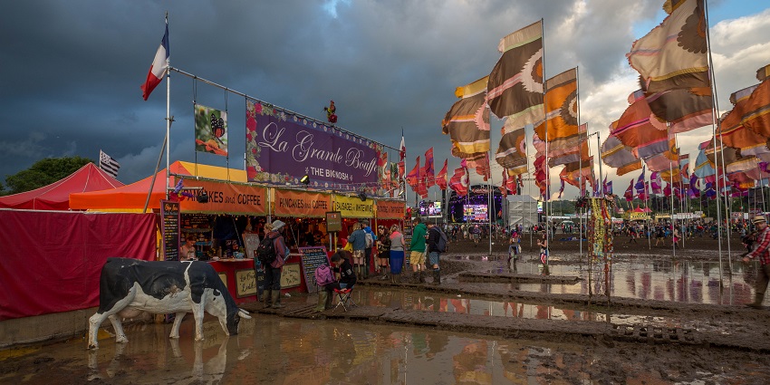 Glastonbury festival with multiple eateries, flags, a cow statue and people in front of the eateries
