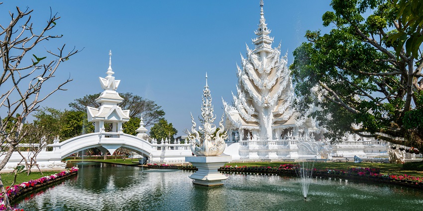 White Temple in Chiang Rai with intricate architecture, pond, fountains, flowers, and trees under the clear blue sky.
