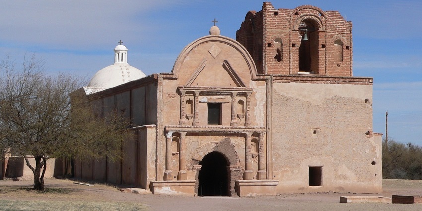 Weathered adobe church walls and arches surrounded by desert plants under a clear sky.