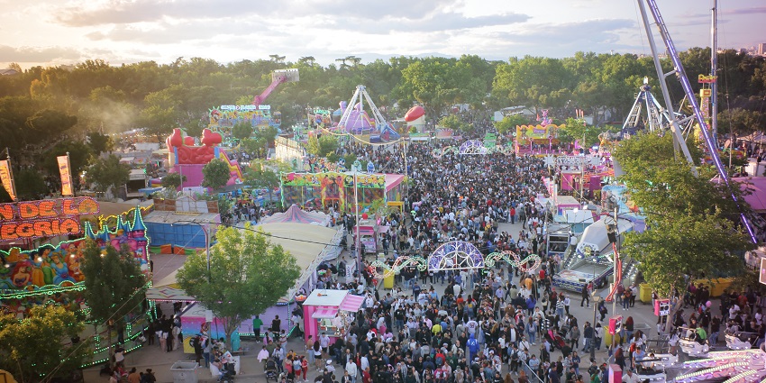 A lively crowd fills the colourful fairground surrounded by rides and game stalls.