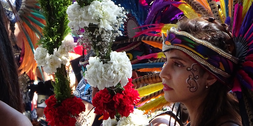 A girl in Aztec attire with a Virgin of Guadalupe image during a vibrant colourful celebration.