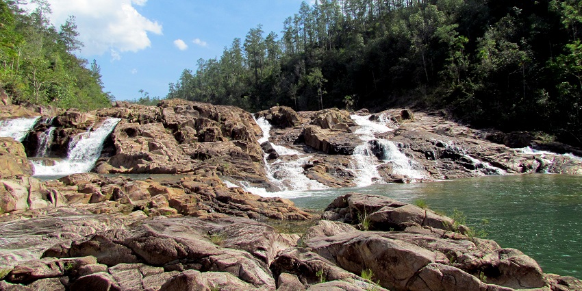 Rocky stream with small waterfalls, natural pools, and forest under a bright blue sky.