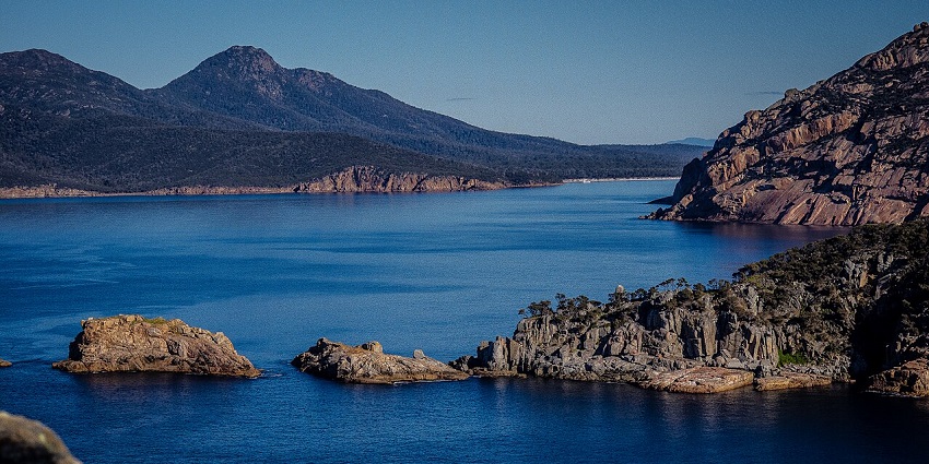 Rocky coastline with deep blue water and rugged mountains under a clear, bright blue sky.