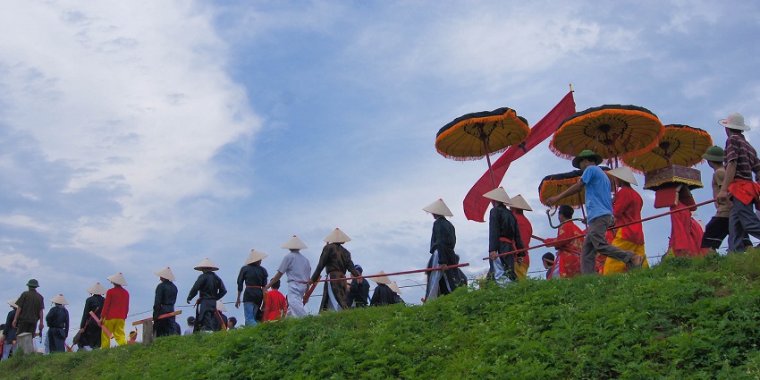 People in traditional attire walking in a ceremonial procession with umbrellas and flags.