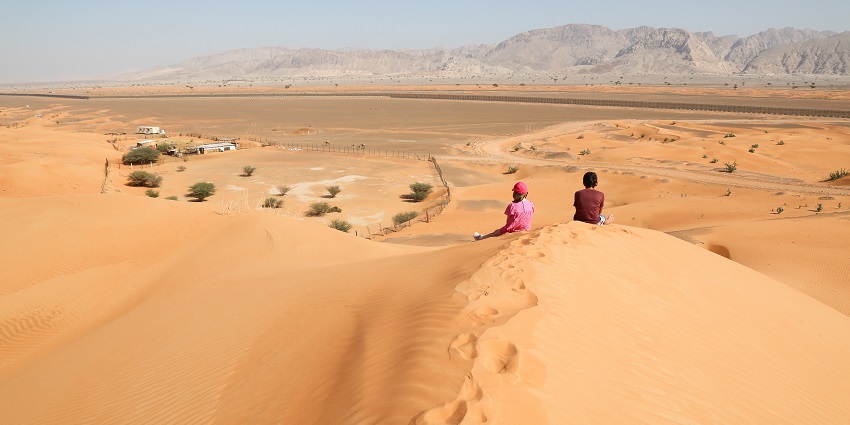 The Hajar Mountains are seen from a vast stretch of desert area with two people admiring their beauty.
