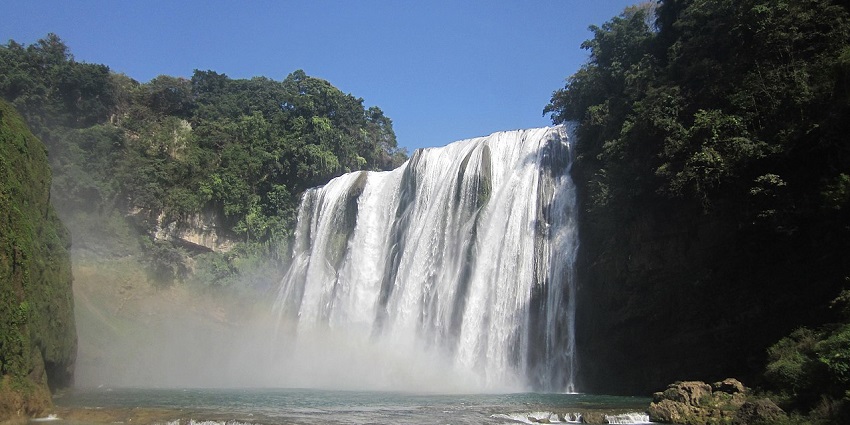 Huangguoshu Waterfall falls dramatically over dark, textured rocks into a foggy basin.