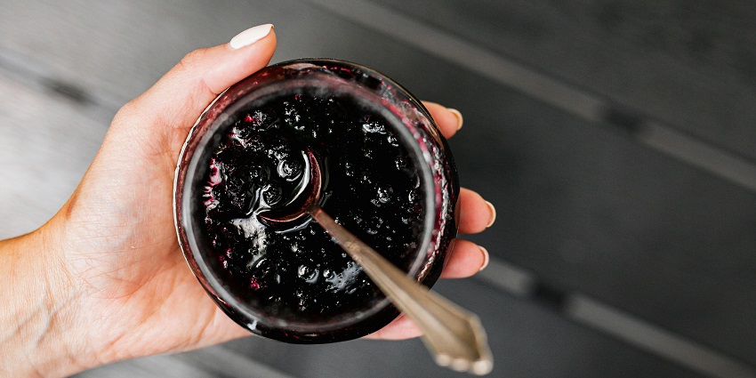 A hand holding a jar of homemade huckleberry jam, showcasing its rich purple color and tempting, glossy texture.