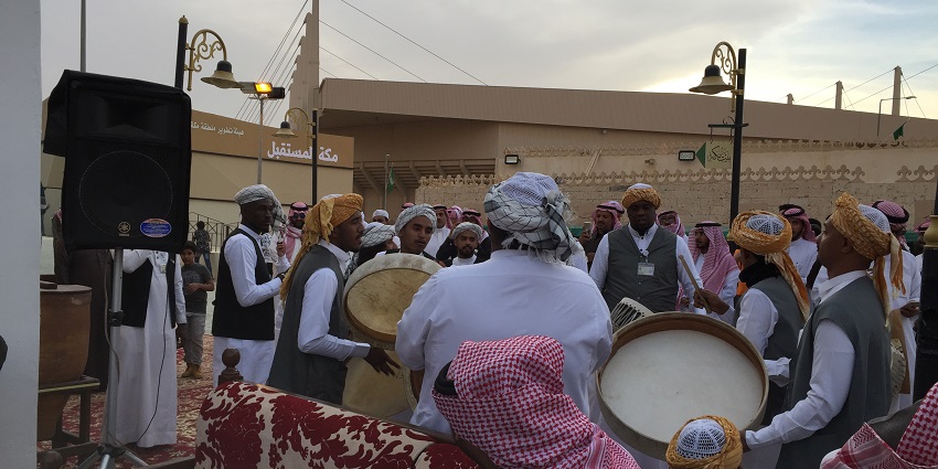 Men in traditional attire celebrate with drums and lively dancing outdoors, and street lamps