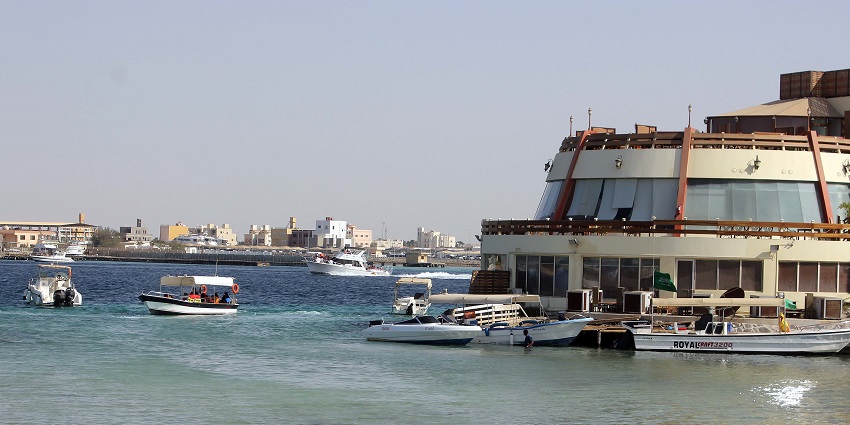 Boats docked by a round waterfront building in Jeddah with the city skyline behind.