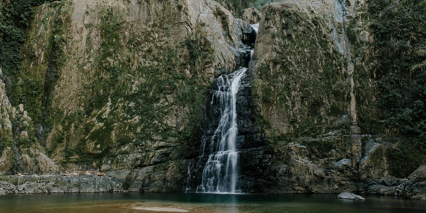A narrow stream of water flows between a moss-covered rocky cliff into a clear pool.