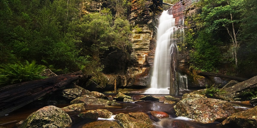 Waterfall flowing over layered rock cliffs into a stream surrounded by mossy boulders.