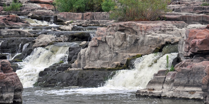 A small waterfall flows through layered rock formations with sparse greenery plants.