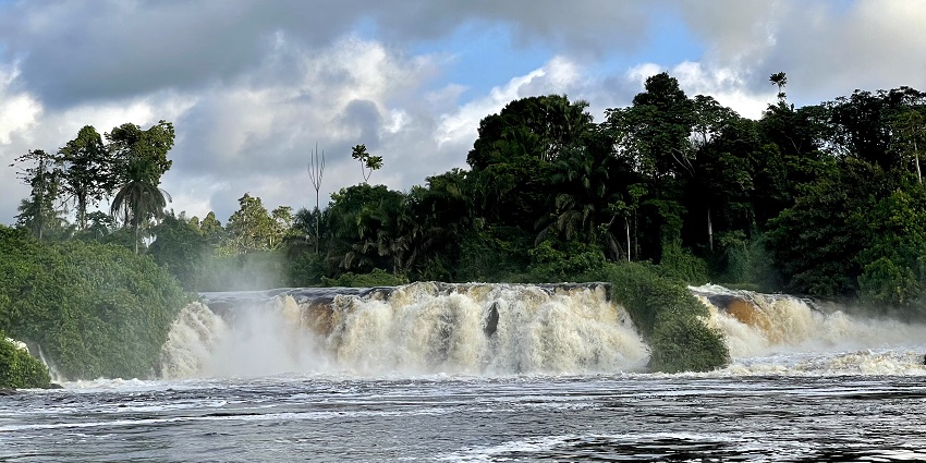 Lobé Falls, Cameroon, cascading under a blue, cloudy sky surrounded by lush green forest.