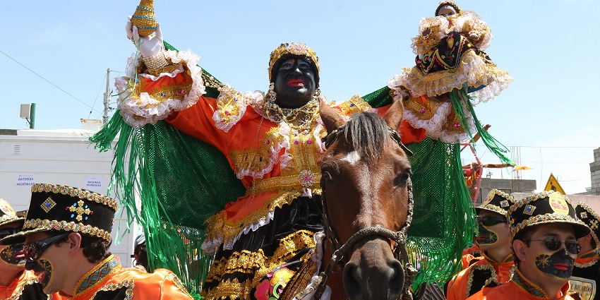 Man in black mask and vibrant costume leads Ecuador’s Mama Negra parade with music, color, and satire.