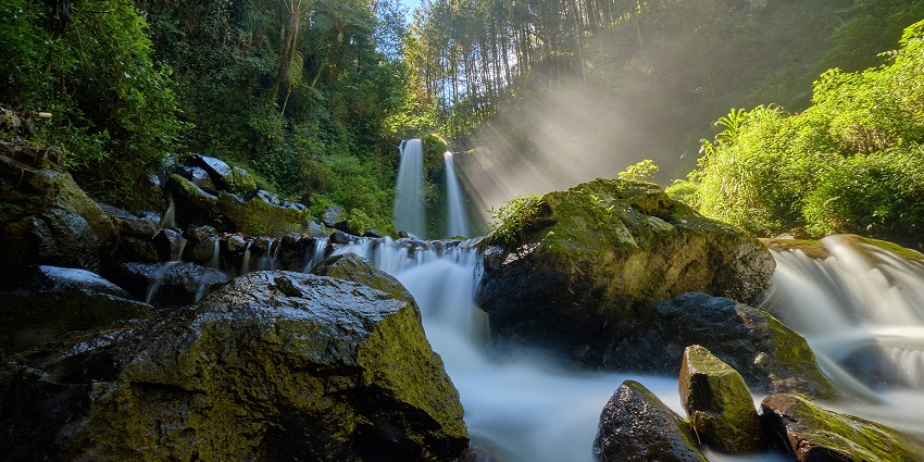 Meloonbua Waterfalls cascade over rocky cliffs into a clear pool, surrounded by lush greenery.