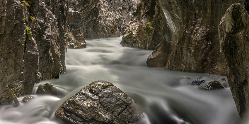Waterfall cascading between grey-black rocks into a cave-like stream, and some green plants.