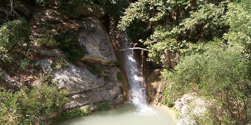 Neda Waterfall flows into a turquoise pool surrounded by limestone cliffs & dense greenery.