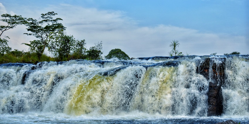 A wide waterfall flows over rocks with trees and a blue sky in the background.