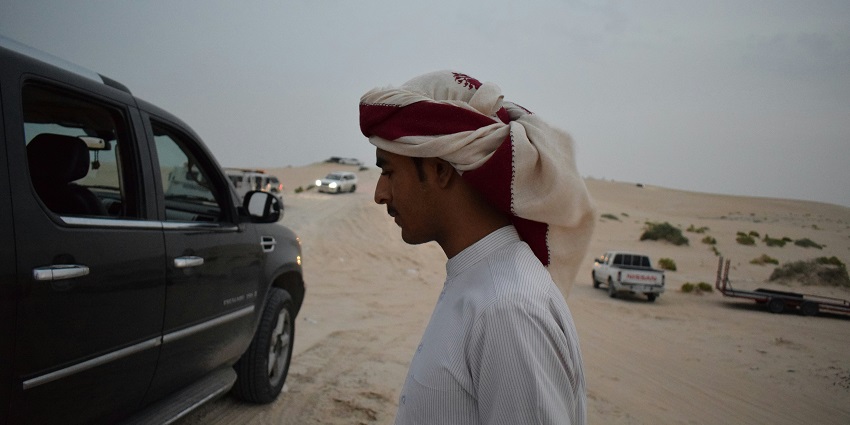 Man in traditional attire at a desert car rally with SUVs and dunes in the background.