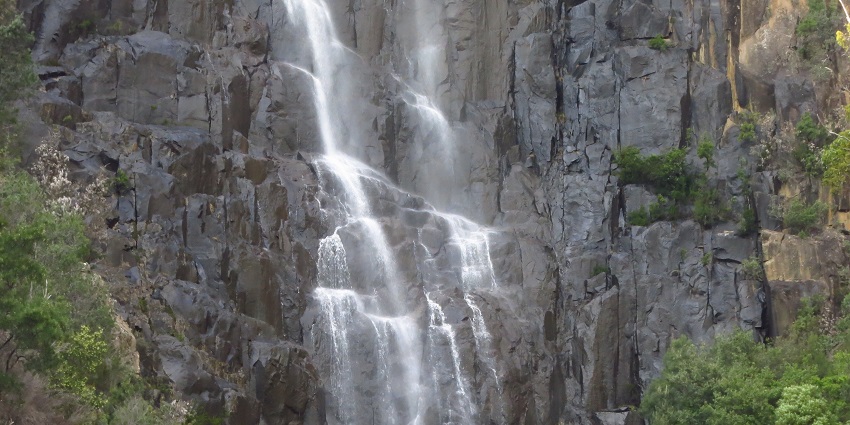 A tall and narrow waterfall tumbling over a rocky cliff, surrounded by trees and plants.