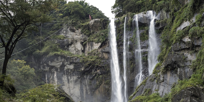 View of Penglai waterfall cascades through layered rocks amid dense green forest and cliffs.