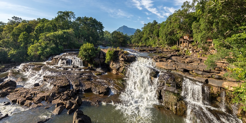 A stunning waterfall cascades down rocks under a clear blue sky with lush greenery around.