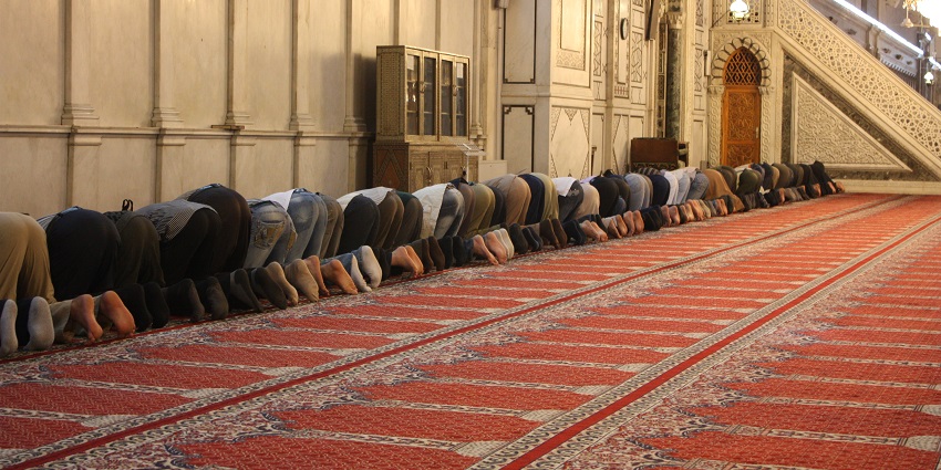 People offer prayers on a red handwoven carpet inside a mosque kneeling and bowing down