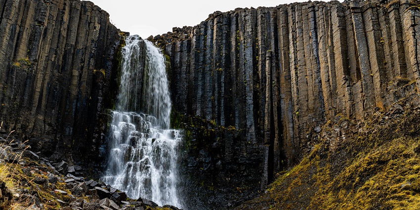 Waterfall cascades down dark basalt columns surrounded by moss-covered rocky slopes.
