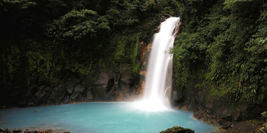 Rio Celeste Waterfall cascades into vibrant turquoise blue water in the waterfalls in Costa Rica’s lush rainforest.