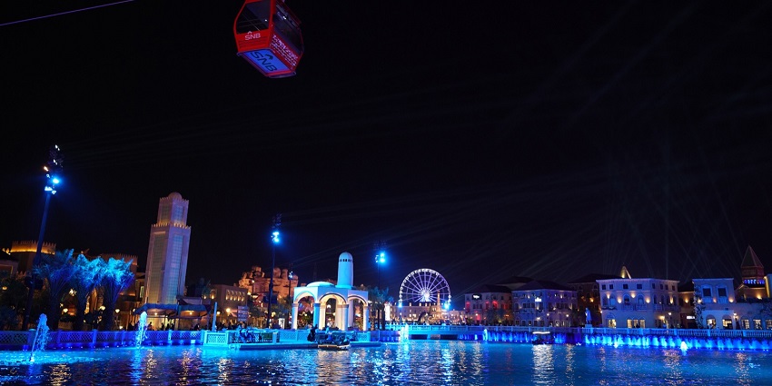 Night view of Riyadh Season with cable car, water, Ferris wheel, and lit-up buildings.