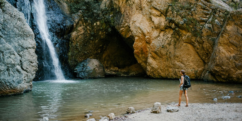 A person with a backpack stands under a narrow waterfall surrounded by huge boulders.