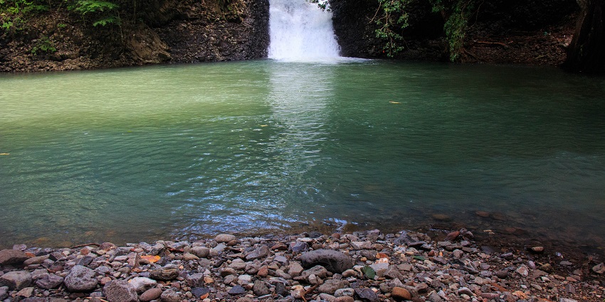 Waterfall cascading down between a muddy cliff into a calm pool surrounded by stones.