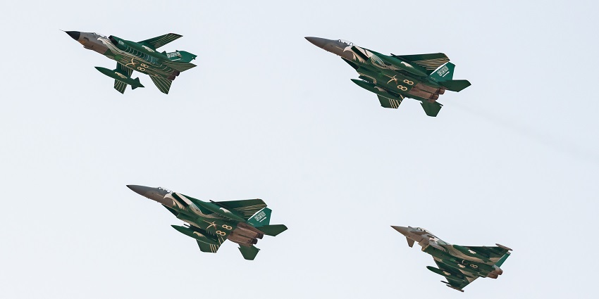 Four green camouflage war planes fly in close formation against a light, cloudless blue sky.