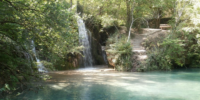 Skra Waterfalls flowing into a vibrant pool surrounded by moss-covered rocks and forest.