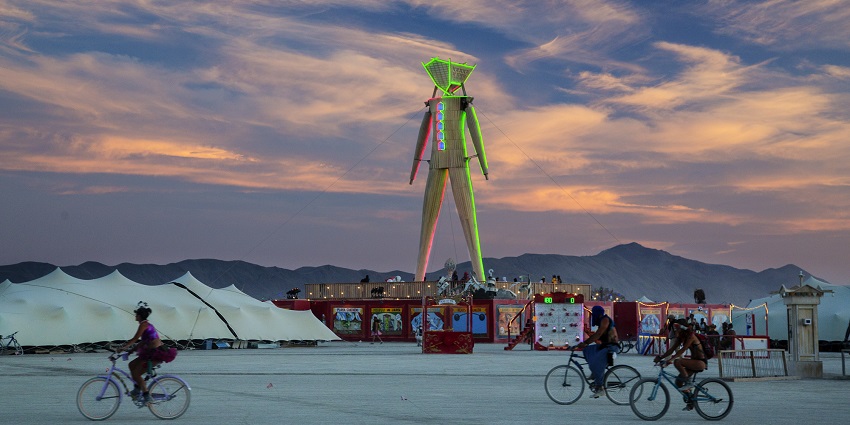 Burning man structure standing tall, people riding bikes and different tents all around