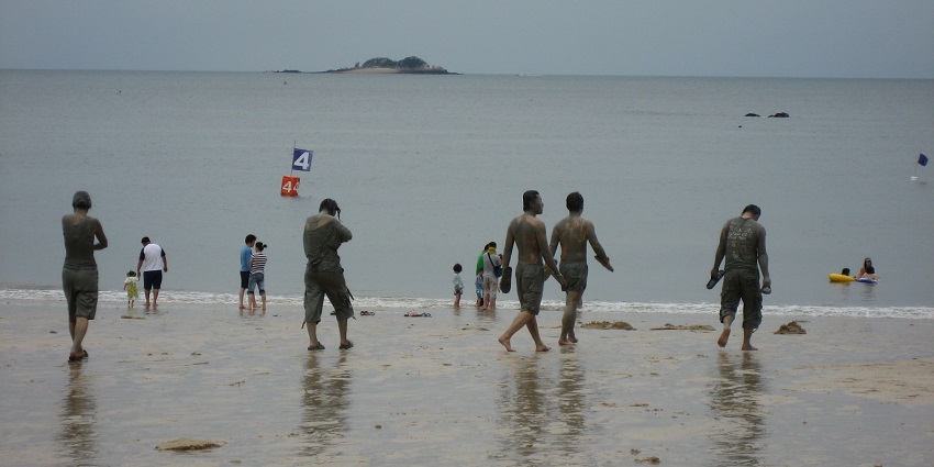 People covered in mud walking on the beach towards the water and others playing