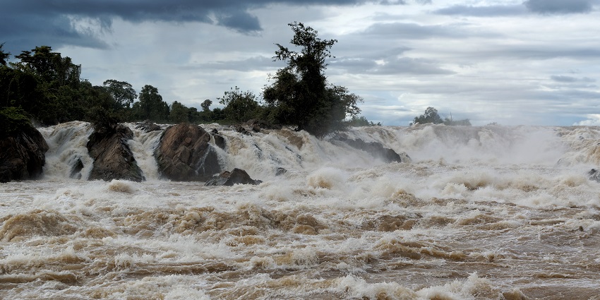 Fierce and muddy waterfall rushing over dark rocks with heavy currents under a cloudy sky