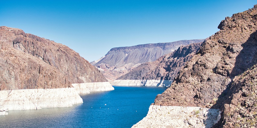 Calm blue lake with boats on the water, surrounded by rocky desert hills and sandy shores.