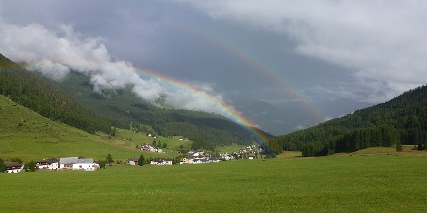 Val Mustair Valley with cloud-covered peaks, distant buildings, and a rainbow in valleys in Switzerland.