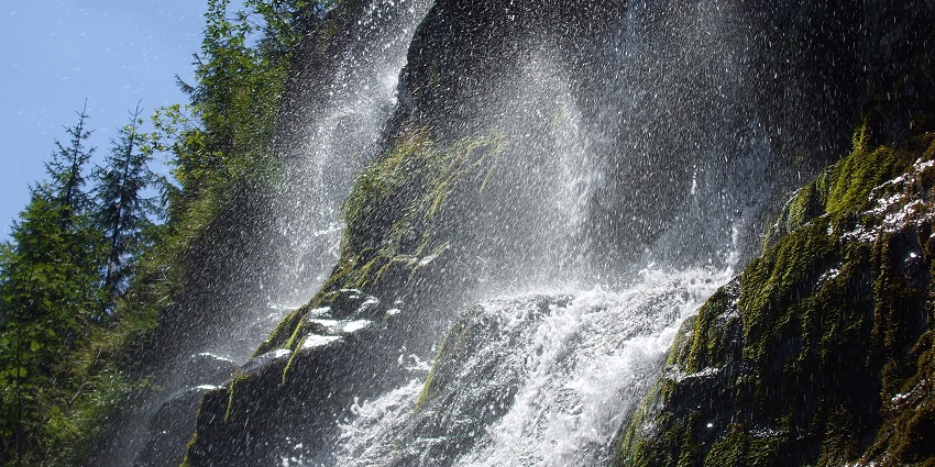 A wide waterfall is streaming across a cliff while clouds are drifting above the forested land.