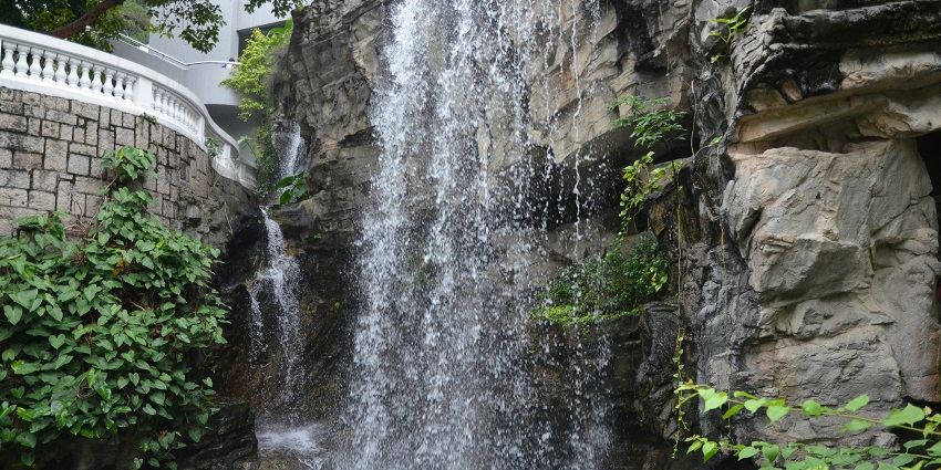 People walk past a serene waterfall surrounded by greenery in a peaceful park in a city.